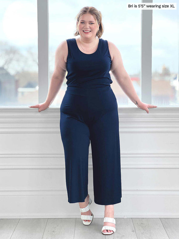 Miik model Bri (five feet five, size extra large) standing in front of a window/white wall wearing Miik's Kimmay open-back capri jumpsuit in navy with white sandals