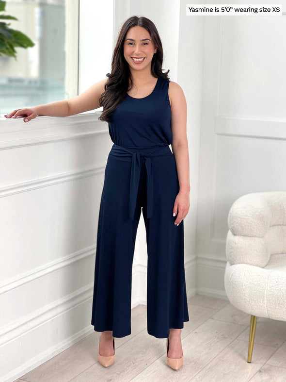 Miik model Yasmine (five feet tall, size extra small, petite) smiling next to a window/white wall wearing Miik's Kimmay open-back capri jumpsuit in navy with high heels