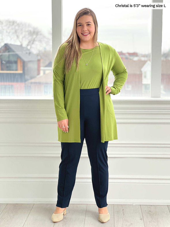 Woman standing in front of a window/white wall wearing Miik's Marcella cardigan with pockets in green moss with a top in the same colour and a navy pant