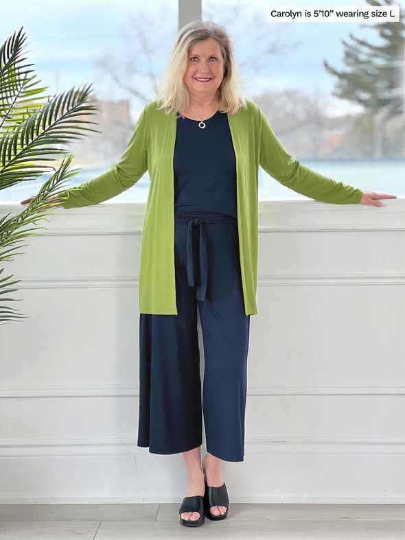 Miik model Carolyn (five feet ten, size large) smiling wearing Miik's Marcella cardigan with pockets in green moss with a navy capri jumpsuit