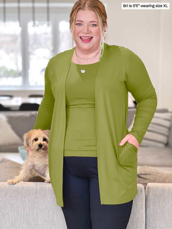 Woman smiling standing in front of a couch next to a dog wearing Miik's Marcella cardigan with pockets in green moss with a matching colour top and navy pant.