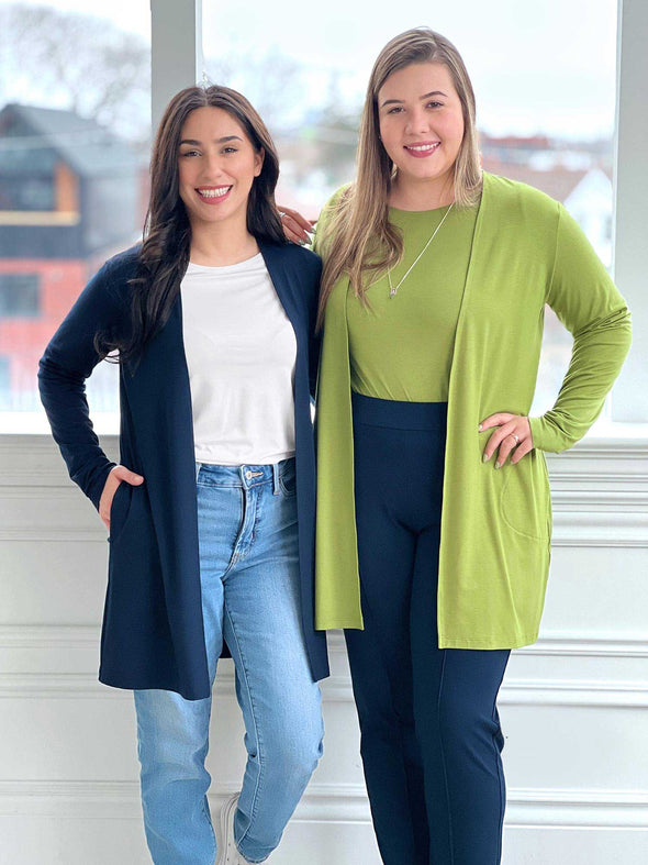 Two women standing next to each other in front of a window wearing both Miik's Marcella cardigan with pockets. On in navy and the other in green moss