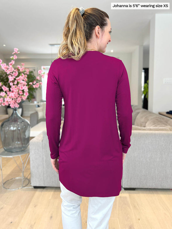 Miik model Johanna (5 foot 6, size extra small) showing the back of the Marcella tunic length cardigan in ruby while looking to the side, smiling, in a high end living room.