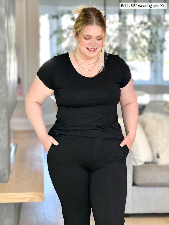 Miik model Bri (five foot five, size extra large) wearing the Marianna classic t-shirt in black with black pants in a living room with her hands in her pockets.
