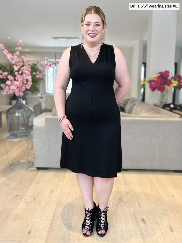 Woman smiling standing in a living room wearing Miik's Mary Jo sleeveless v-neck dress in black with a matching colour high heels