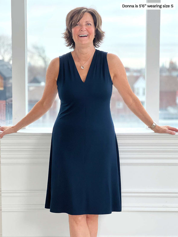 Woman smiling standing in front of a window/white wall wearing Miik's Mary Jo sleeveless v-neck dress in navy