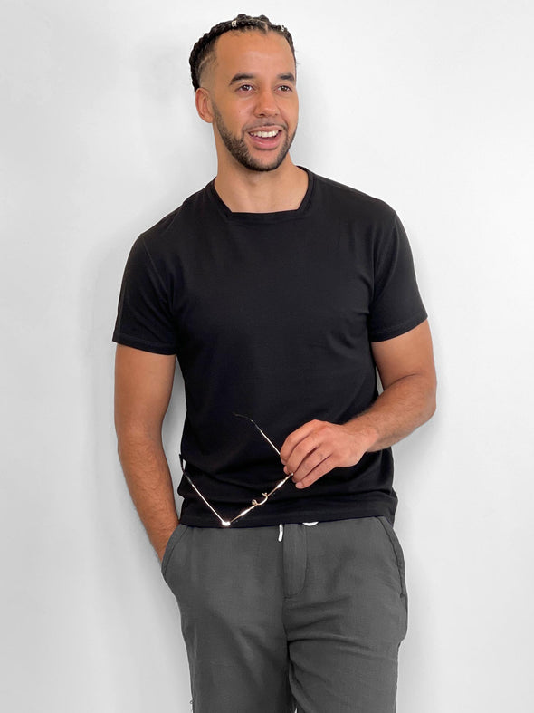 Man standing in front of a wall wearing Miik's Mick square neck t-shirt in black with grey pants.