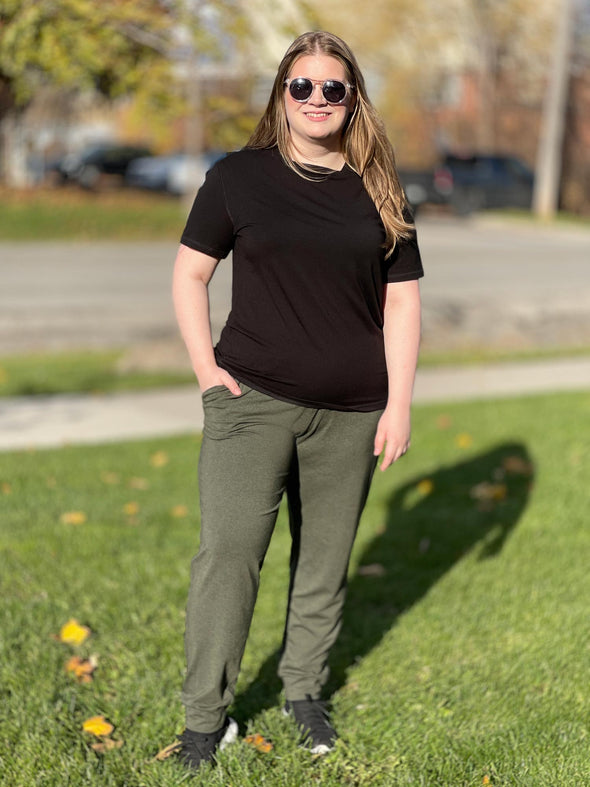 Woman standing on grass wearing Miik's Mick square neck t-shirt in black with green sweatpants.