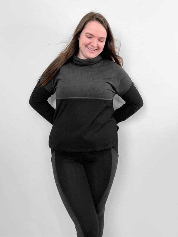 Woman standing in front of a wall wearing Miik's Oaklynn colour block cowl top in grey with matching leggings.