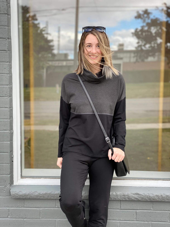 Woman standing in front of a window wearing Miik's Oaklynn colour block cowl top in grey with black pants.