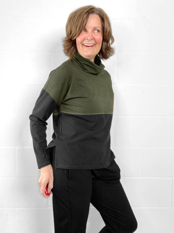 Woman standing in front of a wall wearing Miik's Oaklynn colour block cowl top in green with black pants.
