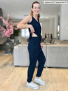 Miik model Johanna (5 foot 6, size extra small) wearing the Perle open-back tank top jumpsuit in a living room, smiling while lifting the back of the jumpsuit's top portion to show that the jumpsuit is only attached at the front.