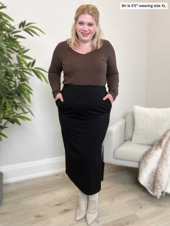 Woman standing in a room wearing Miik's Priya modern long sleeve v-neck top in brown chocolate melange along with a midi skirt in black