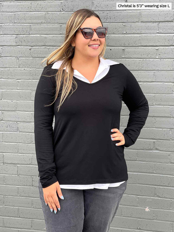 Woman standing in front of a wall wearing Miik's Priya modern long sleeve V-neck top in black on top of a white collared shirt with grey jeans.