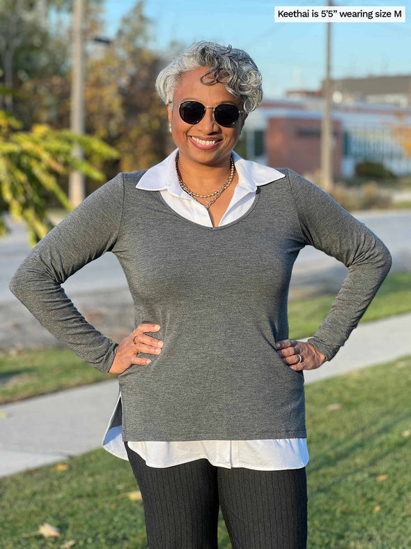 Woman standing next to a sidewalk wearing Miik's Priya modern long sleeve V-neck top in granite grey over a white collar shirt with grey pants.