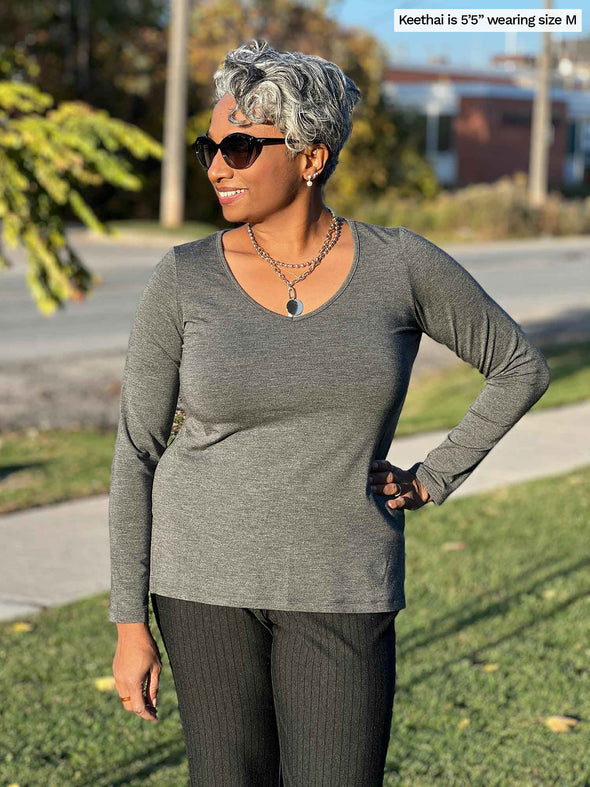 Woman standing by the sidewalk wearing Miik's Priya modern long sleeve V-neck top in granite grey with grey pants.