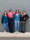 Group of women standing in front of a wall wearing Miik's Priya modern long sleeve V-neck top in teal, teal melange, pink pomegranate and black with jeans.