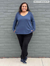 Woman standing in front of a wall wearing Miik's Priya modern long sleeve V-neck top in navy melange with black pants. 