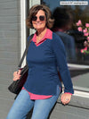 Woman leaning against a window wearing Miik's Priya modern long sleeve V-neck top in navy melange on top of a pink collared shirt with blue jeans.