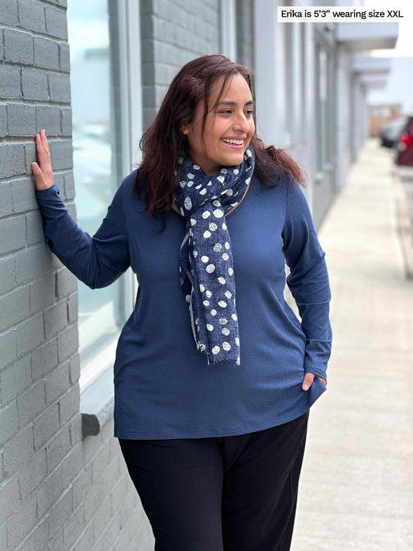 Woman standing next to a wall wearing Miik's Priya modern long sleeve V-neck top in navy melange with black pants and a blue scarf around her neck.