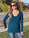 Woman standing next to a sidewalk wearing Miik's Priya modern long sleeve V-neck top in teal with jeans.