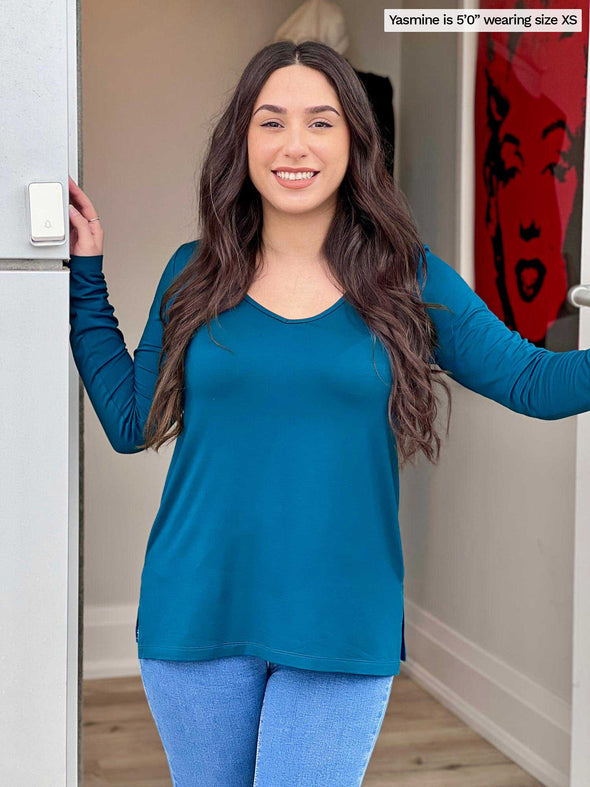 Woman standing at a doorway wearing Miik's Priya modern long sleeve V-neck top in teal with blue jeans.