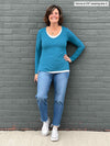 Woman standing in front of a wall wearing Miik's Priya modern long sleeve V-neck top in teal melange with jeans.
