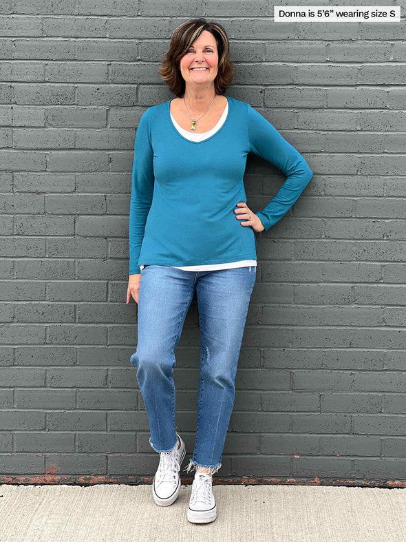 Woman standing in front of a wall wearing Miik's Priya modern long sleeve V-neck top in teal melange with jeans.