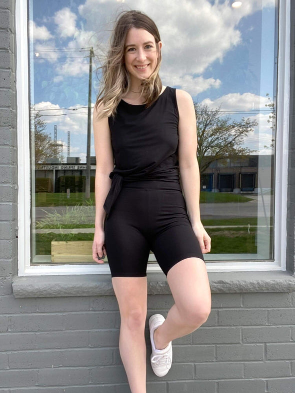 Woman smiling and looking away while leaning against a window wearing Miik's Raven high waisted biker shorts in black with a matching colour tank top 