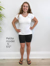 Woman standing in front of a white wall wearing Miik's Raven biker shorts in black and a white t-shirt. The image has also description for petite model