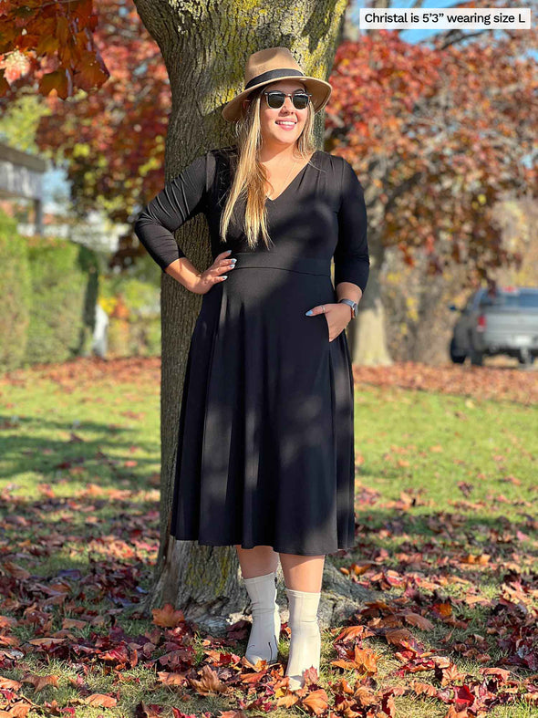Woman smiling while leaning against to a tree wearing Miik's Rebel midi flounce dresss with pockets in black with boots, a hat and sunglasses 