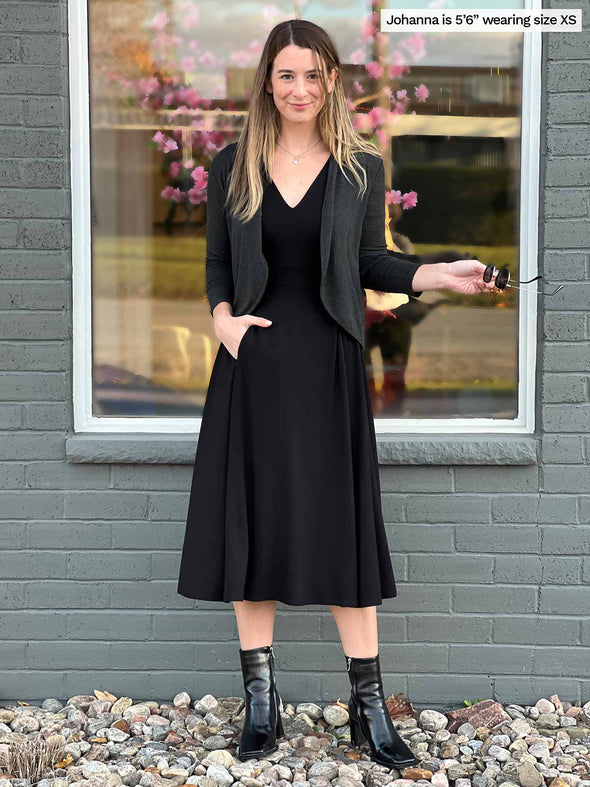 Woman standing in front of a window wearing Miik's Rebel midi flounce dress with pockets in black with a cropped cardigan in charcoal and black boots