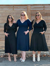 Three women standing in front of a wood wall wearing Miik's Rebel midi flounce dress with pockets in the two different colours available: navy and black