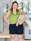 Miik model Christal (five feet three, size large) smiling while holding a puppy and hands on pockets wearing Miik's Reesa racerback high-low tank top in green moss with navy dressy short