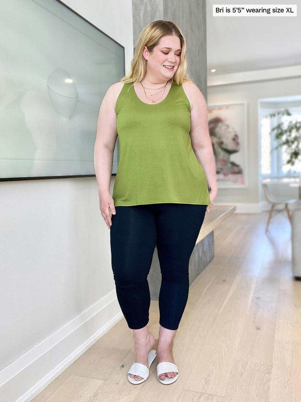 Miik model Bri (five feet five, size extra large) standing in a living room and looking down wearing a capri legging with Miik's Reesa racerback high-low tank top in green moss