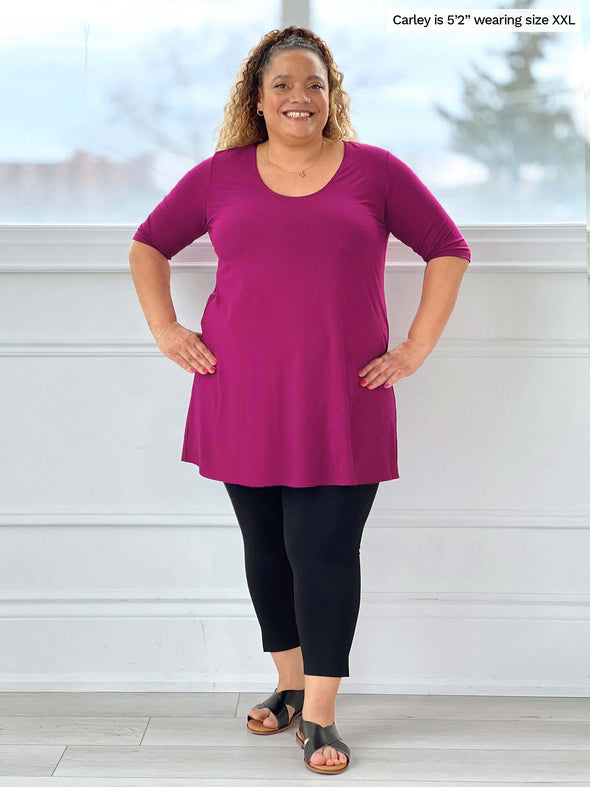 Miik model Carley (five feet two, size duble extra large) smiling wearing Miik's Rocelle half-sleeve scoop neck tunic in ruby with a capri legging in black
