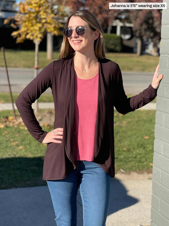 Woman standing next to a wall wearing Miik's Rory waterfall cardigan in brown over a pink pomegranate top with blue jeans. 