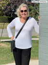 Woman standing next to a brick wall wearing Miik's Sarvi v-neck long sleeve top in white with black pants, sunglasses and a crossbody purse