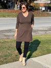 Woman smiling and lookind down wearing Miik's Saya tunic with pockets in chocolate melange, black leggings, boots and sunglasses 
