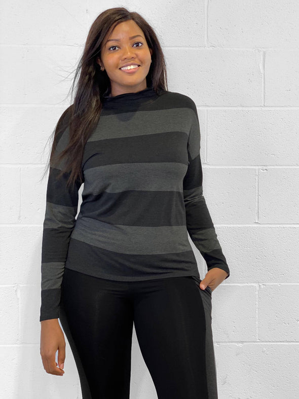 Woman standing in front of a white wall wearing Miik's Scarlett mock neck sweater in dark ash stripe with a black legging