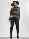 Woman standing in front of a white wall wearing Miik's Scarlett mock neck sweater in dark ash stripe along with a black lounge jogger, boots, a black purse and a big hat 