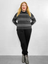 Woman smiling standing in front of a white wall wearing Miik's Scarlett mock neck sweater in dark ash stripe with black jogger and boots 