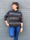Woman standing in front of a brick wall and looking away wearing Miik's Scarlett mock neck sweater in dark ash stripe with jeans and her hand on her hip  