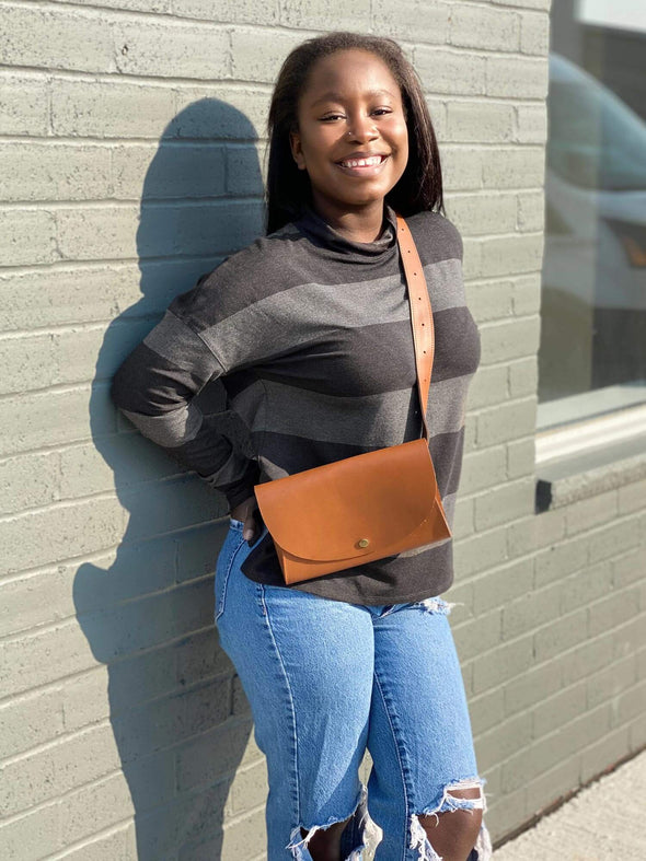 Woman leaning against to a brick wall wearing Miik's Scarlett mock neck sweater in dark ash stripe with jeans and a crossbody purse