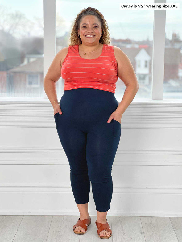 Miik model Carley (five feet two, size double extra large) smiling with hands on pockets wearing Miik's Seana high waisted pocket capri legging in navy with a striped papaya tank top 