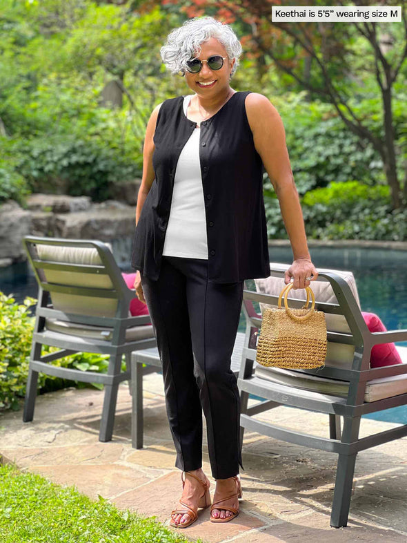 Woman leaning against chairs in front of a pool smiling wearing a white top with Miik's Shane reversible button-up top opened in black with matching black pintuck dress pants.