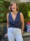 Woman standing in front of outdoor couch laughing wearing Miik's Shane reversible button-up top in navy stripe with first button opened and folded in and pairing it with white pants.