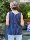 Woman standing in front of outdoor couch with her back towards the camera showing the back of Miik's Shane reversible button-up top in navy with the buttons showing, pairing it with white pants.