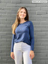 Woman smiling in front of a brick wall wearing Miik's Shannon long sleeve reversible tee in navy melange with white jeans