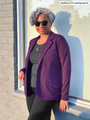 Woman leaning against a window smiling while wearing Miik's Sienna girlfriend blazer in purple colour and a grey top underneath and black pants.